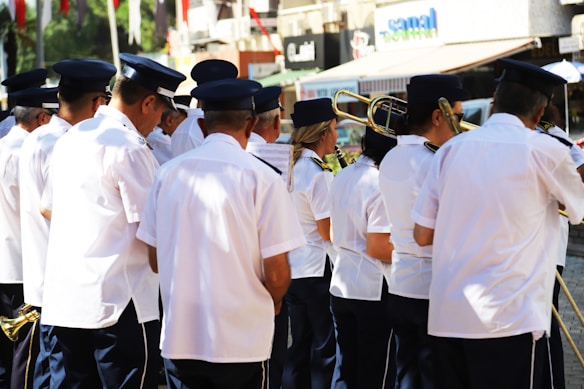 A group of people dressed in matching white uniforms and blue hats are gathered together. They appear to be part of a marching band, holding musical instruments such as brass instruments. The setting seems to be outdoors, possibly on a street, with shop signs and colorful flags visible in the background.