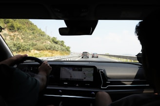 The scene captures the interior view of a moving car on a highway, looking out through the windshield. The driver is steering while a passenger sits beside, and the dashboard displays a GPS map. The road ahead shows a few vehicles, with greenery and hills on the left.