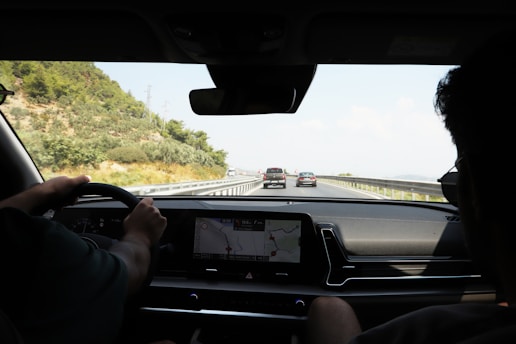 The scene captures the interior view of a moving car on a highway, looking out through the windshield. The driver is steering while a passenger sits beside, and the dashboard displays a GPS map. The road ahead shows a few vehicles, with greenery and hills on the left.