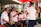 A group of musicians in uniform, likely members of a marching band or musical ensemble, gather under a red canopy. They are playing brass and woodwind instruments, wearing white shirts and blue caps. The setting appears to be outdoors near a modern building with some greenery.