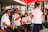 A group of musicians in uniform, likely members of a marching band or musical ensemble, gather under a red canopy. They are playing brass and woodwind instruments, wearing white shirts and blue caps. The setting appears to be outdoors near a modern building with some greenery.