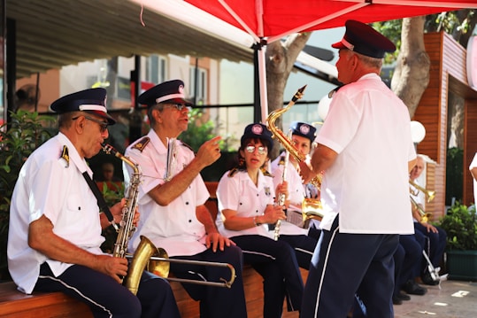 A group of musicians in uniform, likely members of a marching band or musical ensemble, gather under a red canopy. They are playing brass and woodwind instruments, wearing white shirts and blue caps. The setting appears to be outdoors near a modern building with some greenery.