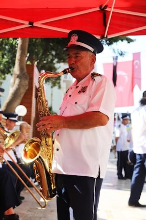 A man wearing a white uniform with a black hat is playing a saxophone under a red canopy. He appears to be part of a band, as there are other musicians in similar uniforms in the background. The setting is outdoors, with flags visible in the background, suggesting a formal or public event.