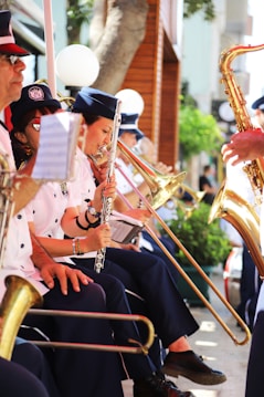 A group of musicians in uniform are playing brass and woodwind instruments outdoors. They appear to be part of a parade or public performance, with focus on individuals playing the trombone, flute, and saxophone. The setting is vibrant and sunny, with trees and a building in the background.