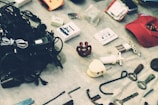 A collection of miscellaneous items displayed on a surface, including tangled cables, a cassette tape, a red cap, and various small tools and objects. The arrangement appears random and the items seem to be of mixed utility and origin.