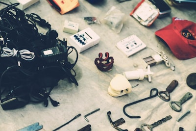 A collection of miscellaneous items displayed on a surface, including tangled cables, a cassette tape, a red cap, and various small tools and objects. The arrangement appears random and the items seem to be of mixed utility and origin.
