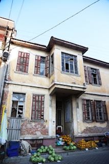 A worn-out, two-story building with peeling yellow paint and several broken windows. The building has wooden shutters painted in a faded red. Below the windows on the ground level, a variety of fruits, including watermelons and pumpkins, are neatly arranged for sale on the pavement.