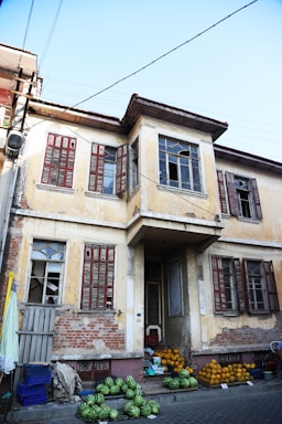 A worn-out, two-story building with peeling yellow paint and several broken windows. The building has wooden shutters painted in a faded red. Below the windows on the ground level, a variety of fruits, including watermelons and pumpkins, are neatly arranged for sale on the pavement.