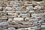 A rustic stone wall composed of variously sized and shaped rocks tightly fitted together. The stones display a mix of earthy colors and textures, with an overall natural and rugged appearance.