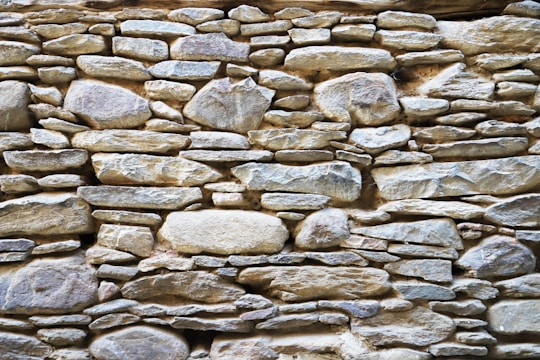 A rustic stone wall composed of variously sized and shaped rocks tightly fitted together. The stones display a mix of earthy colors and textures, with an overall natural and rugged appearance.
