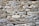 A rustic stone wall composed of variously sized and shaped rocks tightly fitted together. The stones display a mix of earthy colors and textures, with an overall natural and rugged appearance.