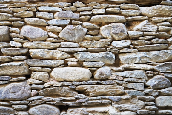 A rustic stone wall composed of variously sized and shaped rocks tightly fitted together. The stones display a mix of earthy colors and textures, with an overall natural and rugged appearance.