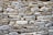 A rustic stone wall composed of variously sized and shaped rocks tightly fitted together. The stones display a mix of earthy colors and textures, with an overall natural and rugged appearance.