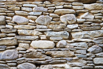 A rustic stone wall composed of variously sized and shaped rocks tightly fitted together. The stones display a mix of earthy colors and textures, with an overall natural and rugged appearance.