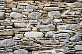 A rustic stone wall composed of variously sized and shaped rocks tightly fitted together. The stones display a mix of earthy colors and textures, with an overall natural and rugged appearance.