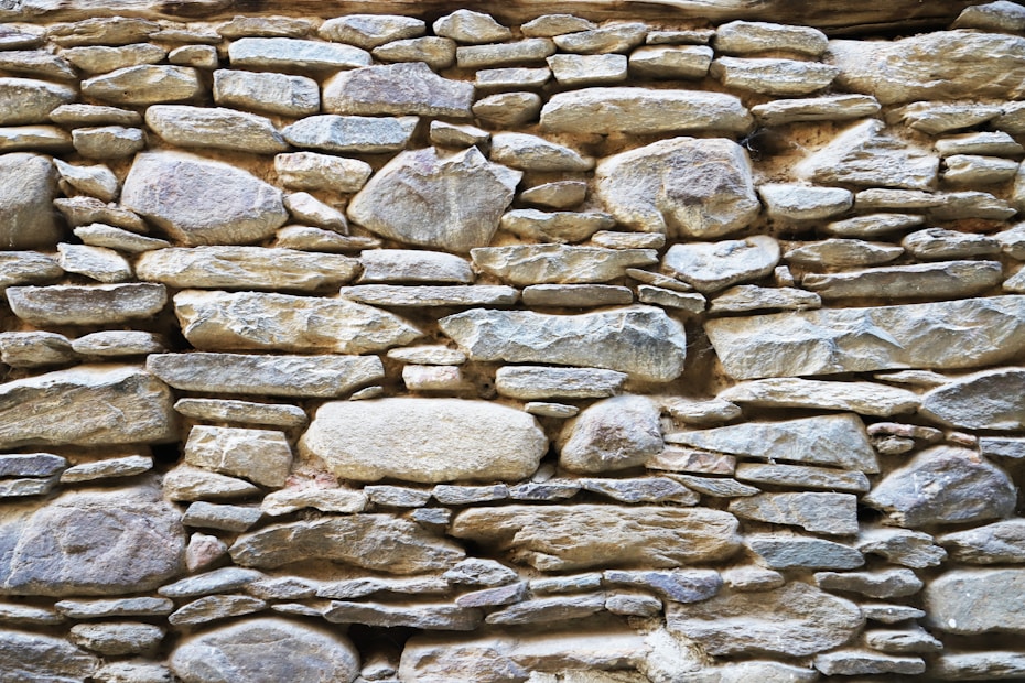 A rustic stone wall composed of variously sized and shaped rocks tightly fitted together. The stones display a mix of earthy colors and textures, with an overall natural and rugged appearance.