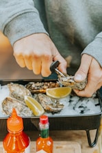 Hands are actively shucking oysters, which are placed on a bed of ice in a tray along with lemon wedges. Two bottles of hot sauce are visible in the foreground, adding a vibrant touch to the scene.