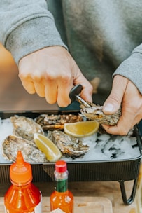 Hands are actively shucking oysters, which are placed on a bed of ice in a tray along with lemon wedges. Two bottles of hot sauce are visible in the foreground, adding a vibrant touch to the scene.