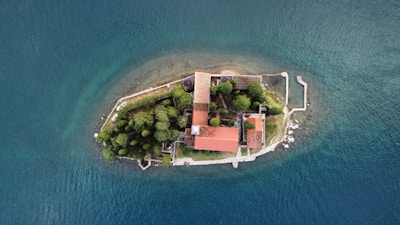 An aerial view of a small island surrounded by water. The island features several buildings with red-tiled roofs nestled among dense green trees. A stone wall lines part of the island's perimeter, with clear blue water visible on all sides.