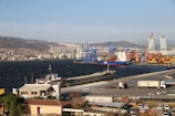 A bustling port scene featuring cranes and a large cargo ship docked near a container terminal. The background includes hills with numerous buildings and a clear blue sky. Several trucks and smaller vehicles are visible on the road adjacent to the port, and modern high-rise buildings can be seen in the distance.