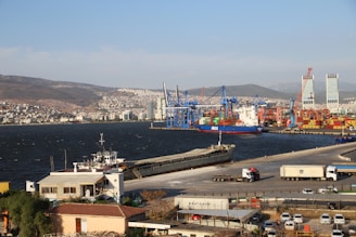 A bustling port scene featuring cranes and a large cargo ship docked near a container terminal. The background includes hills with numerous buildings and a clear blue sky. Several trucks and smaller vehicles are visible on the road adjacent to the port, and modern high-rise buildings can be seen in the distance.