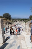A group of tourists walks along an ancient stone path lined with columns and ruins. The scene is set outdoors on a sunny day with a clear blue sky. Vegetation and trees are visible in the background, providing a contrast to the historical architecture.