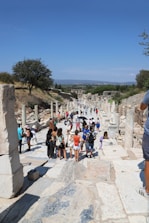 A group of tourists walks along an ancient stone path lined with columns and ruins. The scene is set outdoors on a sunny day with a clear blue sky. Vegetation and trees are visible in the background, providing a contrast to the historical architecture.