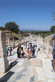 A group of tourists walks along an ancient stone path lined with columns and ruins. The scene is set outdoors on a sunny day with a clear blue sky. Vegetation and trees are visible in the background, providing a contrast to the historical architecture.
