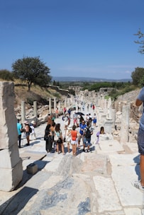 A group of tourists walks along an ancient stone path lined with columns and ruins. The scene is set outdoors on a sunny day with a clear blue sky. Vegetation and trees are visible in the background, providing a contrast to the historical architecture.