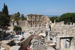 Ancient ruins with numerous stone columns and structures, surrounded by trees and a distant hilly landscape. A group of people are walking and exploring the area, suggesting a historical site visit.