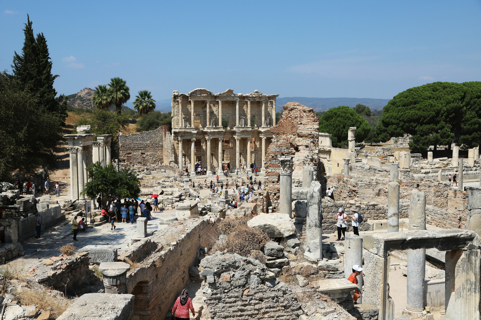 Ancient ruins with numerous stone columns and structures, surrounded by trees and a distant hilly landscape. A group of people are walking and exploring the area, suggesting a historical site visit.