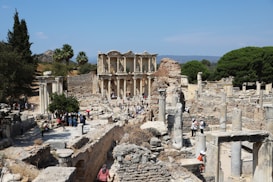 Ancient ruins with numerous stone columns and structures, surrounded by trees and a distant hilly landscape. A group of people are walking and exploring the area, suggesting a historical site visit.
