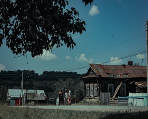 A rural scene featuring an old, rustic wooden house with a weathered roof. The sky is blue with a few clouds. In the foreground, three people are gathered near the road, with one using a wheelchair. A large tree branch partially frames the scene. Power lines stretch across the image.
