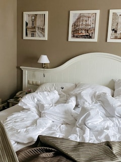 A cozy bedroom features a neatly designed headboard against a light brown wall. Three framed pictures displaying architectural scenes hang above the bed, which is unmade with white, fluffy bedding. A wall-mounted lamp and a bedside table with a telephone add to the decor.
