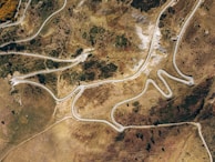 An aerial view of a winding road that navigates through a rugged, mountainous landscape. The terrain features a mix of dry, rocky patches and sparse vegetation, with various shades of brown and green. The road curves sharply, suggesting a challenging and scenic route. There are a few vehicles on the road, indicating activity.