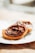Two pastries are placed on a decorative napkin on a wooden surface, each topped with a generous swirl of chocolate icing. The pastries have a golden-brown, crispy texture and are captured in bright, natural light that enhances their glossy appearance.