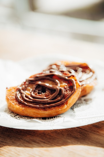 Two pastries are placed on a decorative napkin on a wooden surface, each topped with a generous swirl of chocolate icing. The pastries have a golden-brown, crispy texture and are captured in bright, natural light that enhances their glossy appearance.