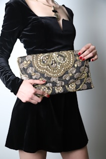A person wearing a black velvet dress holds an intricately designed clutch embellished with gold and silver embroidery. The person's fingernails are painted red to match the elegant style. The clutch features ornate patterns with beads and sequins, adding a luxurious touch.
