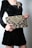 A person wearing a black velvet dress holds an intricately designed clutch embellished with gold and silver embroidery. The person's fingernails are painted red to match the elegant style. The clutch features ornate patterns with beads and sequins, adding a luxurious touch.