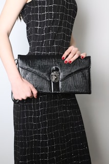 A person is holding a large black leather clutch with a textured surface and a metal clasp. The person is wearing a black dress with a silver grid pattern. The nails are painted red, complementing the elegant and sophisticated look.