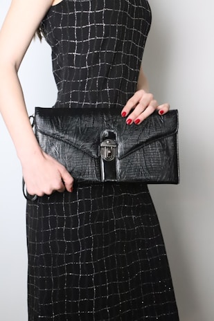 A person is holding a large black leather clutch with a textured surface and a metal clasp. The person is wearing a black dress with a silver grid pattern. The nails are painted red, complementing the elegant and sophisticated look.