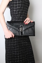 A person is holding a large black leather clutch with a textured surface and a metal clasp. The person is wearing a black dress with a silver grid pattern. The nails are painted red, complementing the elegant and sophisticated look.