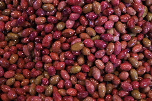 A close-up view of a large quantity of olives, displaying a variety of colors such as red, pink, and green. The olives have a glossy texture, suggesting they are either in brine or oil.