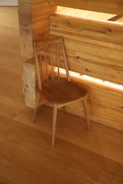 A simple wooden chair with a smooth finish is positioned against a light wooden wall and floor. The chair has a minimalist design with a curved seat and a backrest formed by slender vertical supports.
