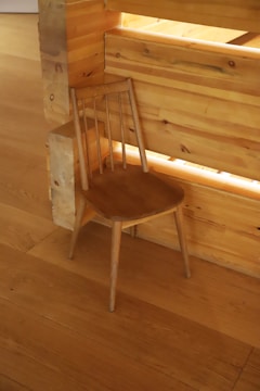 A simple wooden chair with a smooth finish is positioned against a light wooden wall and floor. The chair has a minimalist design with a curved seat and a backrest formed by slender vertical supports.