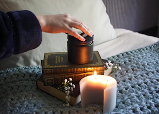 a person lighting a candle with books and a cozy blanket