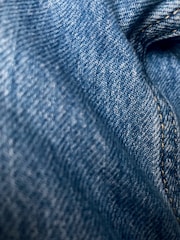 Detailed close-up of blue denim fabric showcasing the texture and weave pattern. The fabric appears slightly wrinkled with visible stitching along one side.