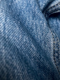 Detailed close-up of blue denim fabric showcasing the texture and weave pattern. The fabric appears slightly wrinkled with visible stitching along one side.