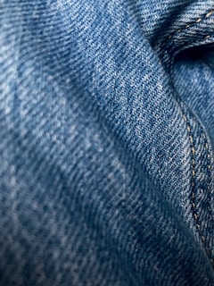 Detailed close-up of blue denim fabric showcasing the texture and weave pattern. The fabric appears slightly wrinkled with visible stitching along one side.