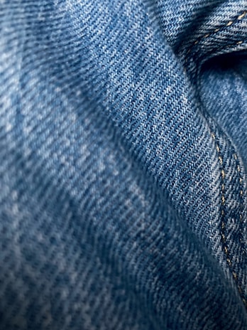 Detailed close-up of blue denim fabric showcasing the texture and weave pattern. The fabric appears slightly wrinkled with visible stitching along one side.